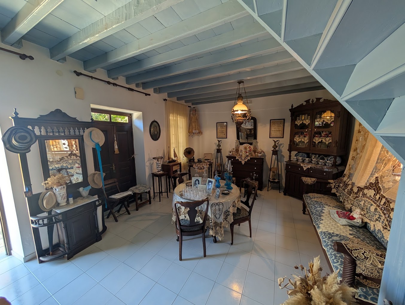 Chalki Trad House Interior - Kolymbia Sea Lines A vintage living room with blue exposed ceiling beams, dark wood cabinets, and a lace-covered dining table set with china and photos.